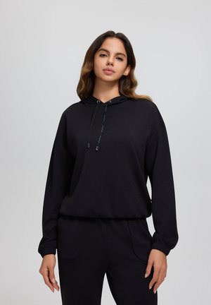 Frau mit langem, welligem Haar trägt schwarzen Kapuzenpullover und passende Hose, stehend vor schlichtem hellgrauem Hintergrund.