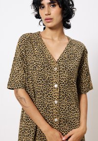 Robe à imprimé léopard avec des manches courtes, un col en V et des boutons en argent. Fabriquée en tissu texturé dans des couleurs beige et noir.