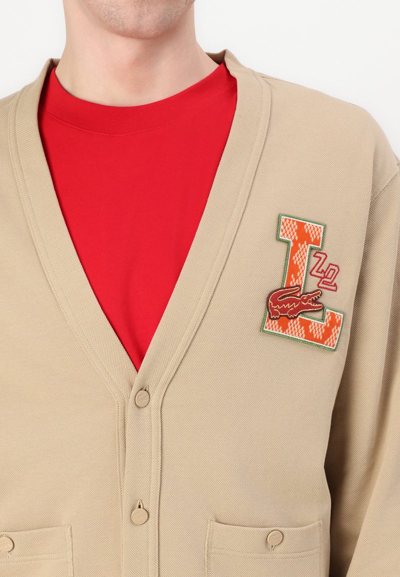 Cardigan en tricot beige avec col en V, boutons sur le devant et deux poches. Présente un 'L' brodé orange et vert ainsi qu'un détail de crocodile. Chemise rouge en dessous.