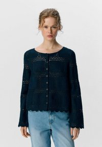 Cardigan en tricot bleu foncé avec motif ajouré, encolure ronde, fermeture à boutons et poignets festonnés. Tissu texturé avec une coupe ample.