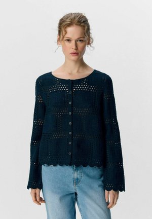 Cardigan en tricot bleu foncé avec motif ajouré, encolure ronde, fermeture à boutons et poignets festonnés. Tissu texturé avec une coupe ample.