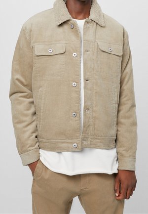 Homme portant une veste en velours côtelé beige avec un col sherpa blanc sur une chemise blanche et un pantalon beige, main dans la poche, debout devant un fond uni.