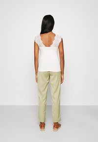 Top blanco con mangas de encaje tipo capa y espalda abierta, combinado con pantalones verde oliva y sandalias marrones de tiras. Fondo neutro.