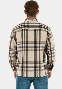 Man som bär blå jeans och en långärmad skjorta i beige, brunt och svart rutig, sedd bakifrån mot en enfärgad bakgrund.