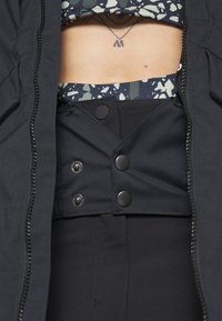Veste zippée noire avec un intérieur ajusté et texturé et des détails à boutons-pression sur un pantalon taille haute noir uni. Couche inférieure avec motif visible.