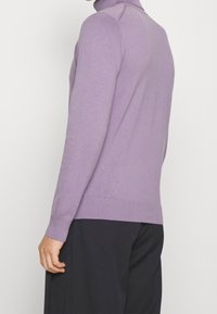 Mann in einem lavendelfarbenen Rollkragenpullover und schwarzen Hosen, von der Seite und von hinten vor einem schlichten Hintergrund gezeigt.