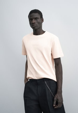 Homme portant une chemise rose clair à manches courtes et un pantalon noir avec une chaîne argentée, debout devant un fond gris clair uni.