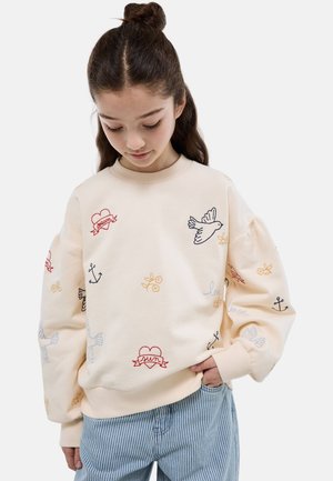 Fille aux cheveux foncés en chignon haut, portant un sweat crème brodé de cœurs, colombes, ancres et fleurs, assorti à un pantalon bleu rayé.