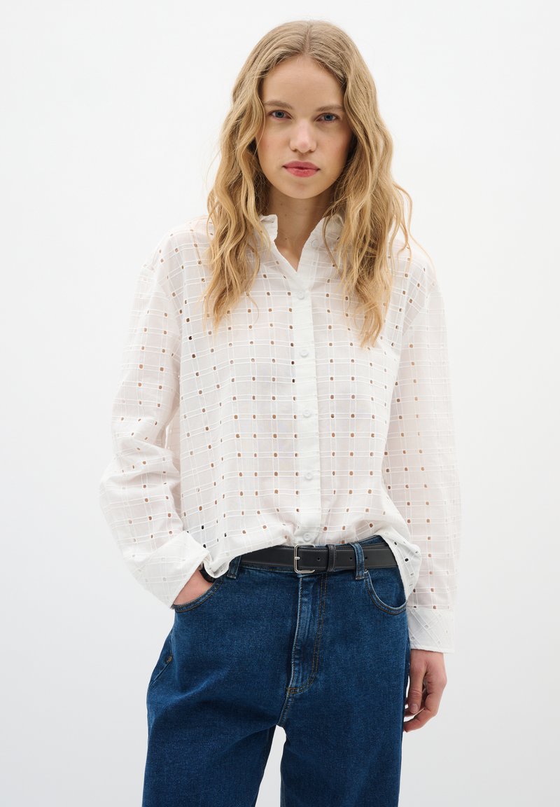 Camicia bianca con bottoni, caratterizzata da un motivo a griglia con piccoli dettagli a intaglio, abbinata a jeans blu a vita alta e una cintura nera.