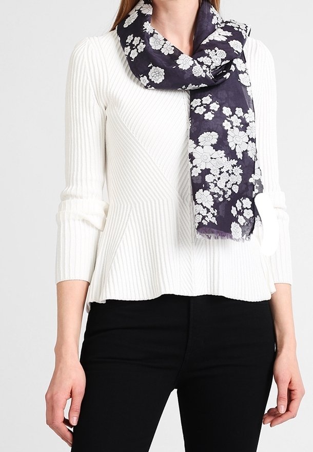 Femme portant un pull en maille côtelée blanc et un pantalon noir, avec une écharpe noire à motif floral blanc drapée autour du cou.