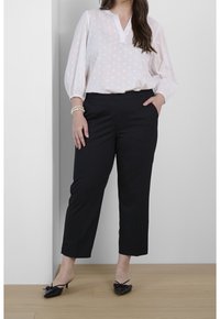 Mujer vestida con blusa blanca texturizada, pantalones negros cortos, tacones negros de punta y pulseras de perlas, de pie contra una pared blanca sobre un suelo de madera.
