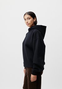 Felpa nera oversize con un grande cappuccio, spalle scese e polsini a coste. Abbinata a pantaloni di velluto a coste marroni.