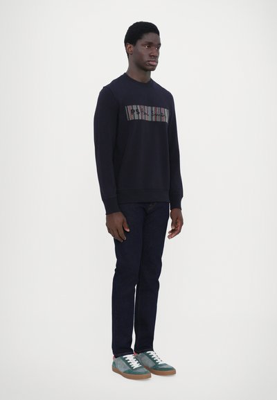Sweatshirt marine avec un motif texturé horizontal sur la poitrine, associé à un jean foncé et des chaussures vertes/grises avec des lacets blancs.