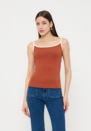 Frau trägt ein rostfarbenes Tanktop mit weißen Besätzen und blaue Jeans, steht vor einem schlichten weißen Hintergrund.