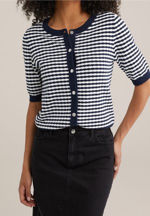 Femme portant un cardigan boutonné à manches courtes bleu marine et blanc à rayures et une jupe en jean noire taille haute.