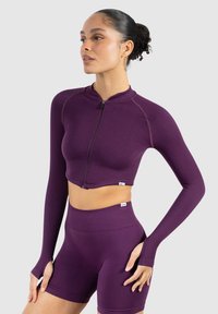 Lilla crop top med lynlås, lange ærmer og tommelfingerhuller, parret med højtaljede lilla shorts. Glat, tætsiddende stof.