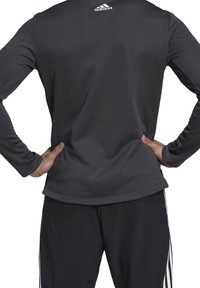 Graues langärmliges Sportoberteil aus feuchtigkeitsableitendem Material mit abgerundetem Saum und dem Adidas-Logo auf dem oberen Rücken. Schwarze Hose mit Streifen.