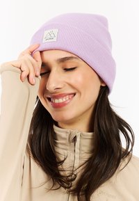 Fialová pruhovaná pletená čepice s logem. Modelka má na sobě hnědou bundu na zip, má dlouhé vlasy, usmívá se a dotýká se své čepice.