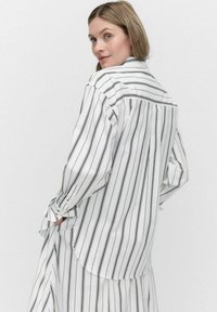 Hvid skjorte med sorte lodrette striber, knap-ned design, lange ærmer, let afslappet pasform og en buet kant bagpå.