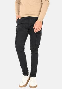 Pantaloni cargo neri con una vestibilità slim, dotati di tasche laterali, una texture liscia e cuciture dettagliate. Abbinati a sneaker bianche e a un maglione beige.