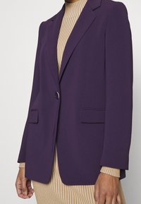 Fioletowa marynarka o dopasowanym kroju, z pojedynczym zapięciem na guzik i dwoma przednimi kieszeniami. Wykonana z gładkiej tkaniny o wyraźnej sylwetce.