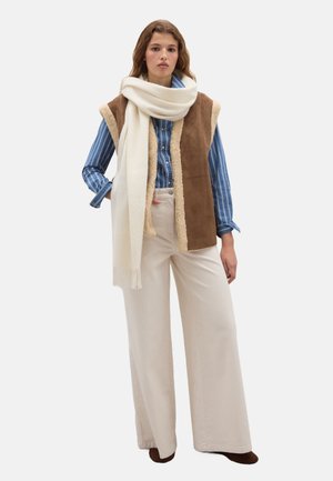 Gilet di shearling marrone indossato sopra una camicia a righe blu, abbinato a pantaloni crema a gamba larga e una spessa sciarpa bianca con dettagli a frange.