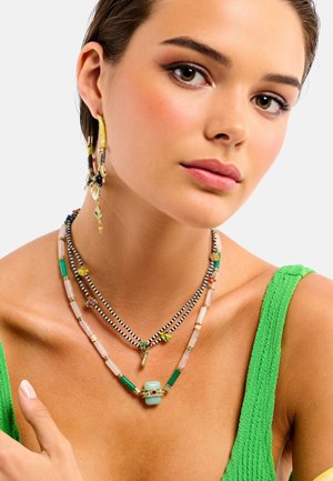 Mehrfarbige, geschichtete Halsketten mit Perlen in Grün, Rosa, Schwarz und Gelb an einem Model, kombiniert mit einem strukturierten grünen Oberteil.