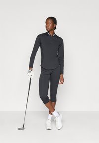 Camiseta deportiva de manga larga negra con cremallera y cuello en contraste, combinada con leggings recortados negros. La modelo sostiene un palo de golf y lleva unos zapatos blancos.