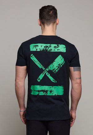 Mann trägt schwarzes T-Shirt mit großem grünem gekreuztem Hackbeil- und Messermotiv auf dem Rücken vor einfarbigem grauem Hintergrund.