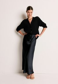 Femeie îmbrăcată cu un cardigan negru cu nasturi, fustă lungă din satin negru, sandale bej și colier din lanț aurit, stând cu mâinile pe șolduri.