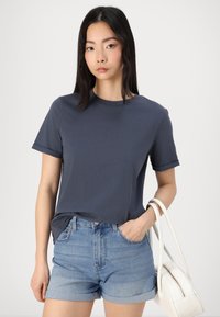 Dark navy short-sleeve t-shirt made of soft fabric, paired with light blue denim shorts. A small white handbag is held in the right hand.