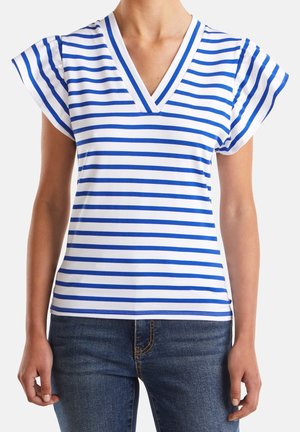 Frau trägt ein weißes V-Ausschnitt T-Shirt mit kurzen Ärmeln und blauen horizontalen Streifen, kombiniert mit dunkelblauen Jeans.