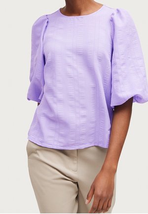 Femme portant un chemisier lilas texturé avec des manches bouffantes jusqu'aux coudes et un pantalon beige sur un fond uni.