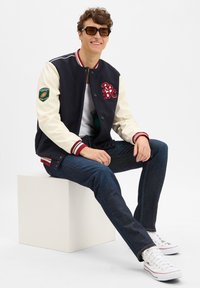 Marineblaues und cremefarbenes College-Jacket mit roten Akzenten, Buchstabenbestickung und einem Aufnäher am Ärmel, kombiniert mit dunklen Jeans und weißen Sneakers.