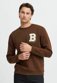 Brun genser laget av mykt stoff, med en kremfarget brodert bokstav "B" på brystet og ribbede cuffs og hem.