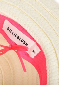 Gros plan sur un chapeau blanc tissé avec une bande intérieure rose portant l’inscription « BILLIEBLUSH 52 » et un ruban rose noué.