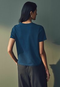 Tmavě modré tričko s krátkým rukávem, hladká tkanina, kulatý výstřih, volný střih, zobrazeno zezadu s šedými kalhotami, minimalistický design.