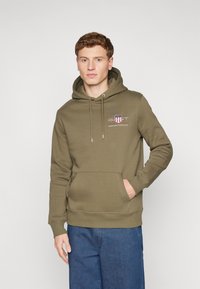 Felpa verde oliva con cappuccio e cordino, tasca frontale e logo stampato. Realizzata in materiale morbido con polsini e orlo a coste. Abbinata a jeans blu.