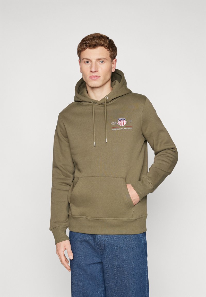 Felpa verde oliva con cappuccio e cordino, tasca frontale e logo stampato. Realizzata in materiale morbido con polsini e orlo a coste. Abbinata a jeans blu.