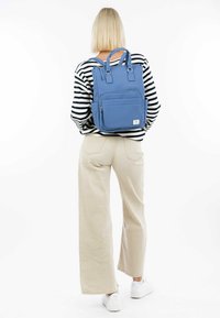 Blue backpack with adjustable straps, front pocket, and top carry handles. Textured fabric, rectangular shape, and white logo tag on the front.