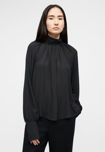 Femme aux longs cheveux bruns portant un chemisier noir ample à col montant avec des manches froncées et des larges poignets, debout avec une main dans la poche.