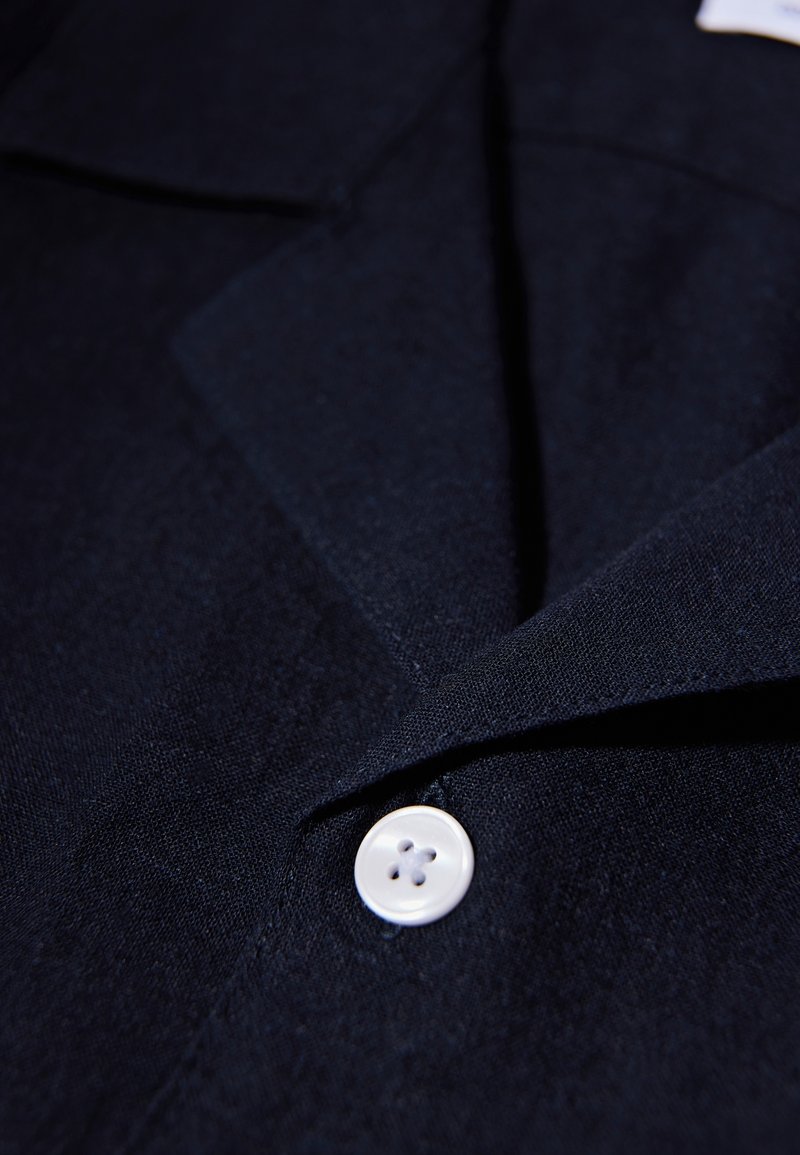 Chemise bleu marine présentant une texture de tissu lisse, un col cranté et un seul bouton blanc à quatre trous.