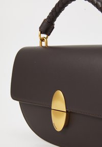 Sac à main en cuir marron foncé avec un fermoir ovale doré, une anse tressée et une texture lisse agrémentée de détails de couture subtils.