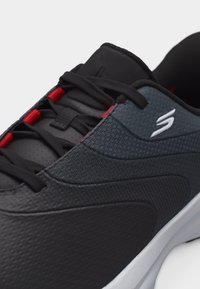 Czarno-grafitowy but sportowy z czerwonymi pętlami na sznurówki, czarnymi sznurówkami, fakturowaną cholewką i białą podeszwą, z widocznym logo Skechers na boku.