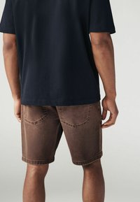 Shorts din denim maro cu o textură ușor deteriorată, având două buzunare la spate și un tiv răsucit, asociați cu o cămașă neagră cu mânecă scurtă.
