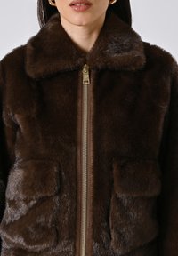 Giacca in finta pelliccia marrone con colletto, zip frontale e due tasche anteriori. Texture morbida e dettagli in metallo lucido.
