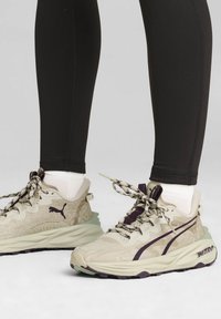 Zapatillas deportivas beige y moradas con tela texturizada, cordones estampados y suelas de goma gruesa. Combinadas con leggins negros.
