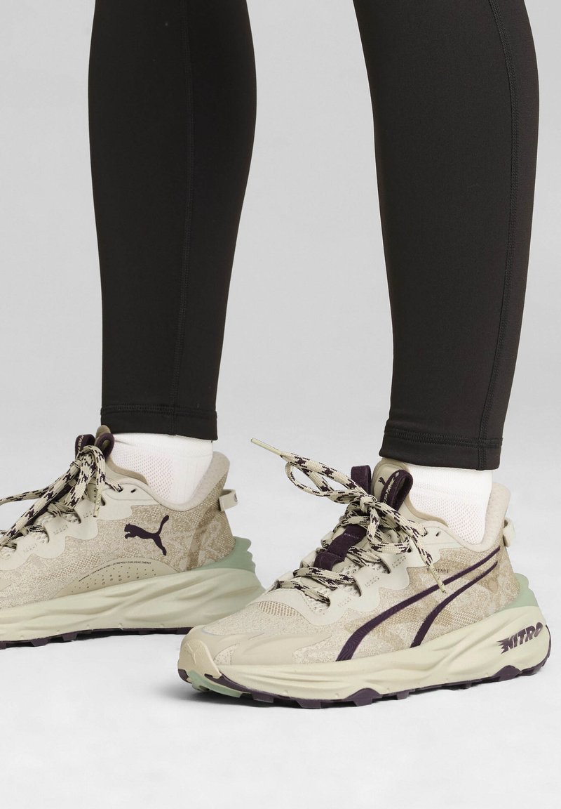 Zapatillas deportivas beige y moradas con tela texturizada, cordones estampados y suelas de goma gruesa. Combinadas con leggins negros.