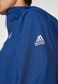 Μπλε ελαφρύ μπουφάν με ψηλό γιακά, κουκούλα, μαύρες λεπτομέρειες, τσέπη με φερμουάρ και λευκό λογότυπο στο δεξί μανίκι. Λείο υφή.