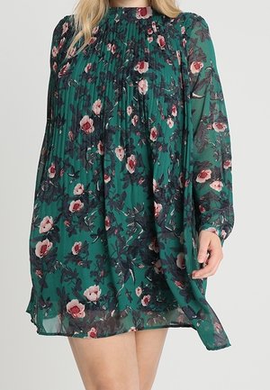 Kvinde iført en grøn blomstret langærmet kjole med plisserede detaljer på brystet og høj hals.